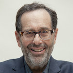 A smiling man with glasses and a beard is wearing a dark blazer against a light background.