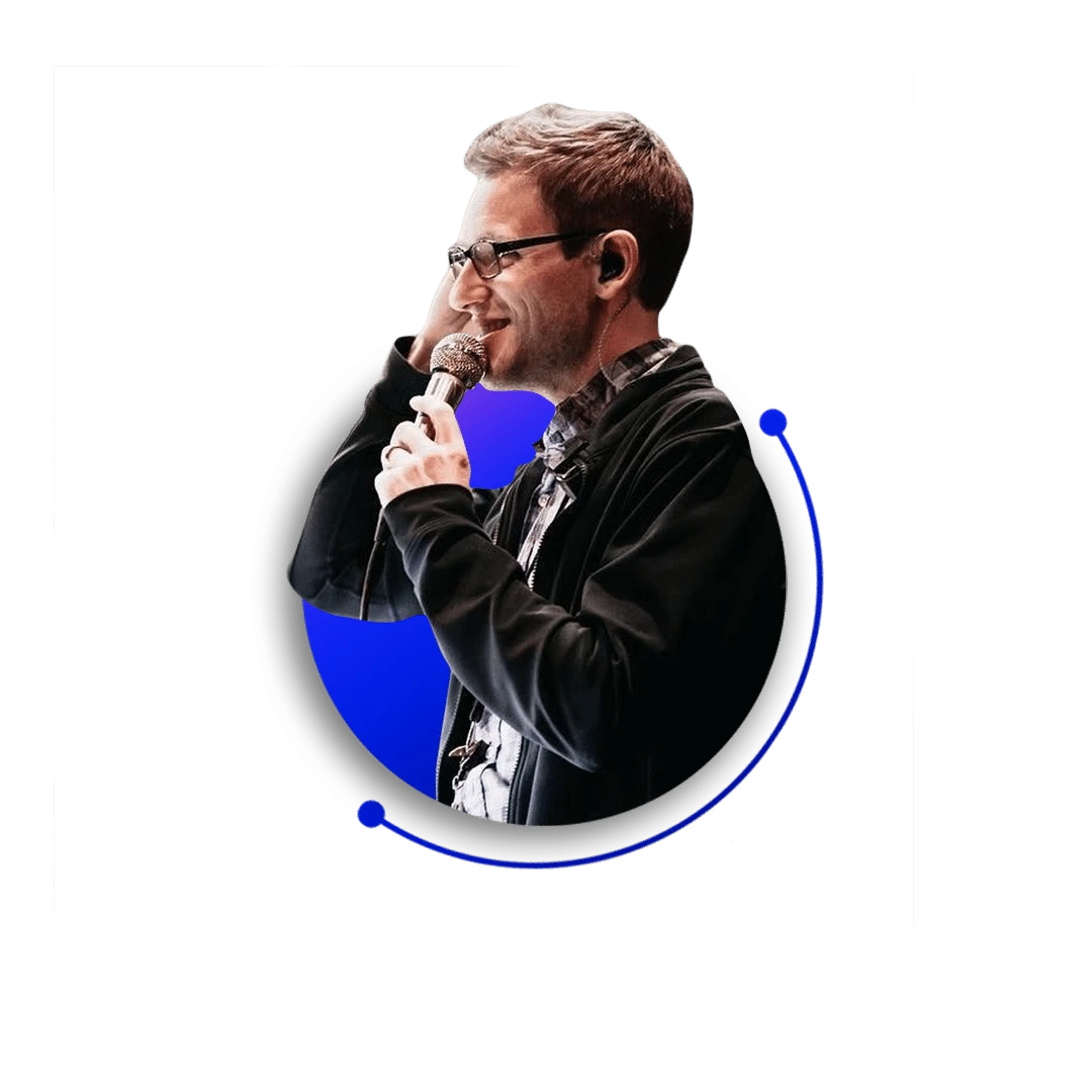 A man, resembling a talking head, wearing glasses, a black jacket, and a patterned shirt holds a microphone to his mouth. He stands against a blue circular background with a modern graphic overlay.