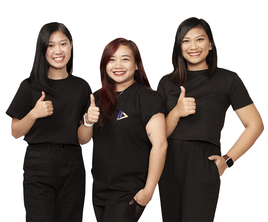 Three people standing against a plain background, all smiling and giving thumbs up. They are wearing black shirts and pants. The person in the center has long red hair, and the others have black hair.