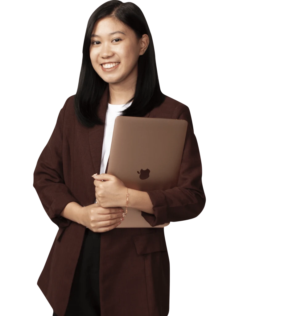 Smiling person with long hair holding a closed laptop, wearing a brown blazer and white shirt, standing against a plain background.