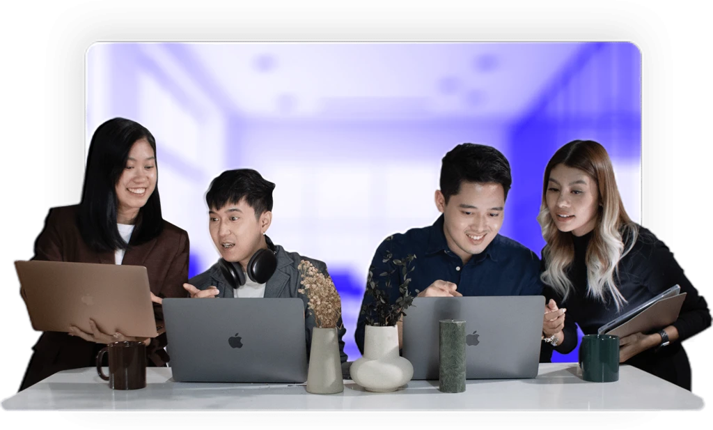 Four people sit around a table working on laptops. They appear engaged and collaborative, surrounded by several vases and cups. A blurred blue background suggests an office environment.