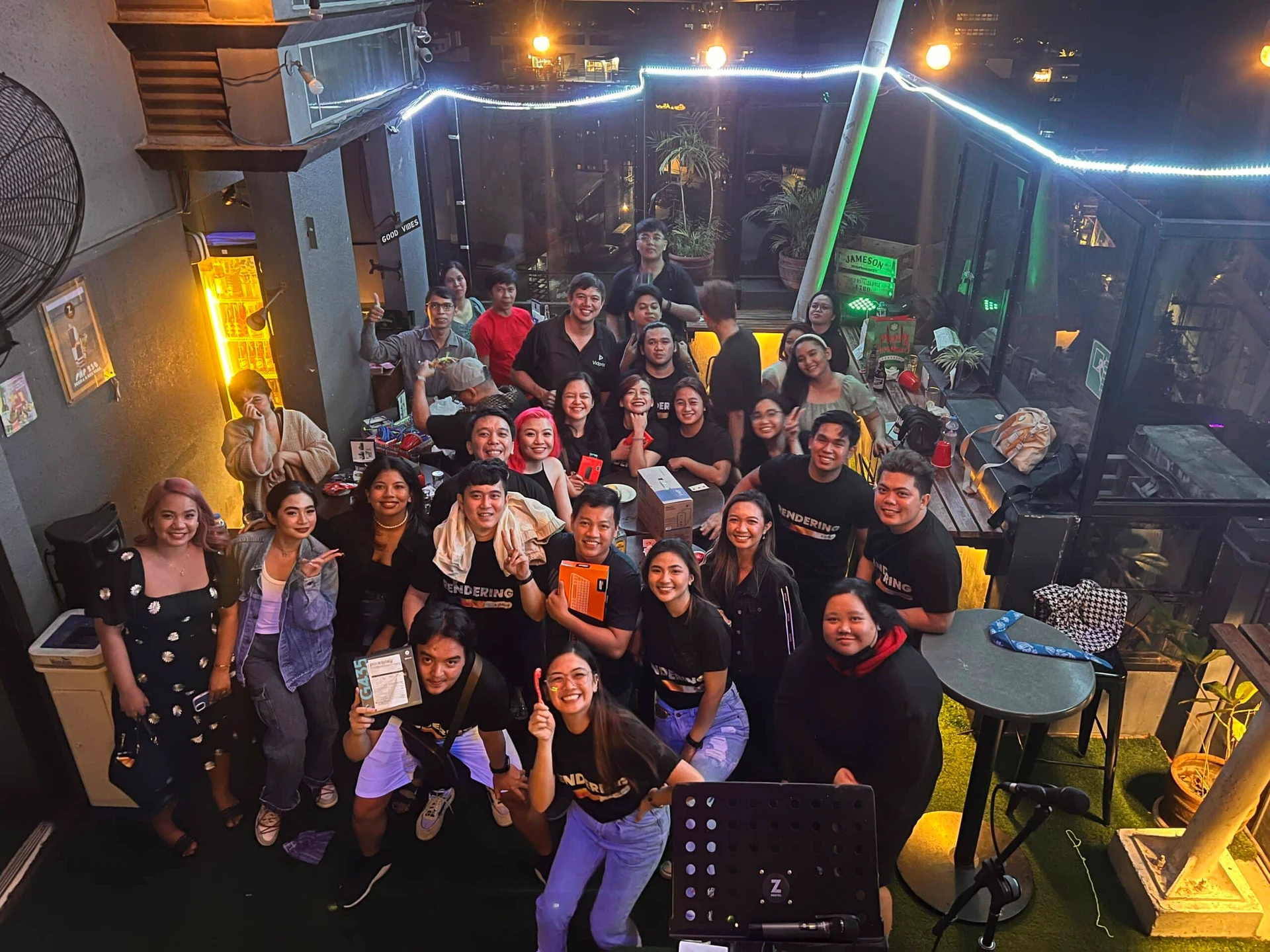 A group of people gathered in a cozy, dimly-lit room, smiling and holding various items. Some are wearing matching t-shirts. The space features plants and string lights, creating a warm atmosphere. Tables with food and drinks are visible in the background.