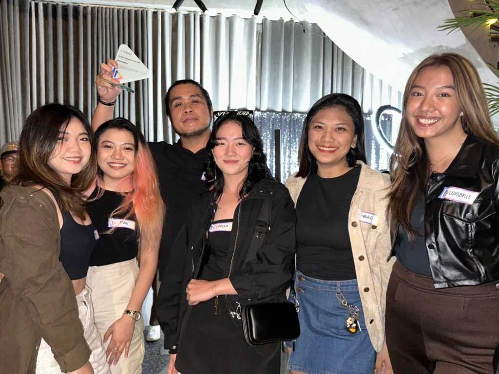 A group of five women and one man smile for a photo in a dimly lit indoor setting. They are dressed in casual, dark-colored outfits. The background features vertical blinds with a shiny, metallic curtain.
