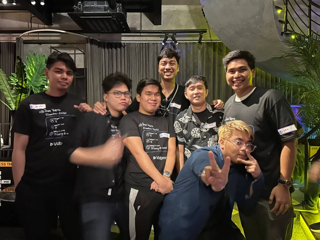 A group of seven young men are posing together indoors, wearing casual attire. Six are in black shirts, and one is making a peace sign. They are smiling and standing in front of a modern, industrial-style wall with plants.