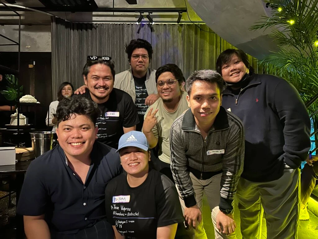 A group of seven people, six men and one woman, smiling and posing together indoors. They are dressed casually and standing in front of a curtain with plants visible in the background.