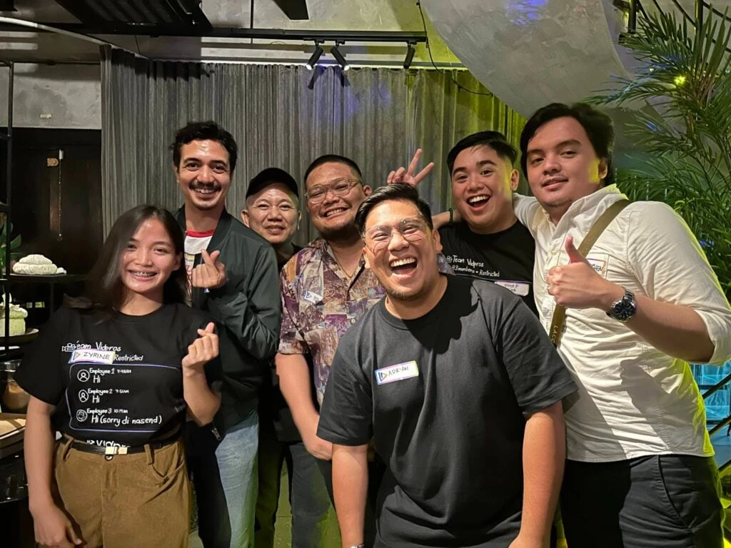 A group of seven people smiling and posing for a photo indoors. They are standing close together, some making playful gestures. The background features a curtain and plants, with warm lighting creating a cheerful atmosphere.