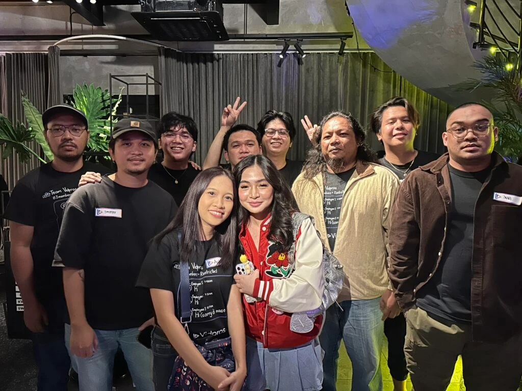 A group of nine people standing together indoors, smiling. They are casually dressed, and two women are at the front, with one wearing a white and red jacket. Some are wearing glasses and hats, and there are plants and curtains in the background.