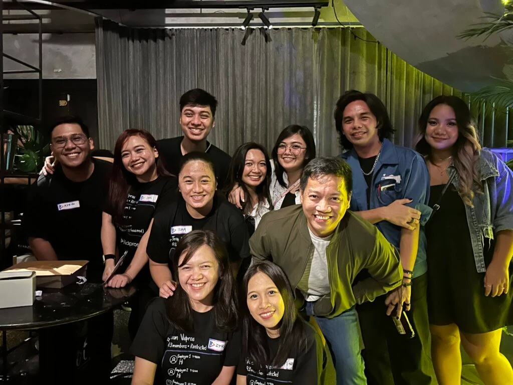 A group of twelve people smiling and posing together indoors. Some are wearing name tags and black shirts. They appear to be at a social gathering or event. The background features curtains and a plant.