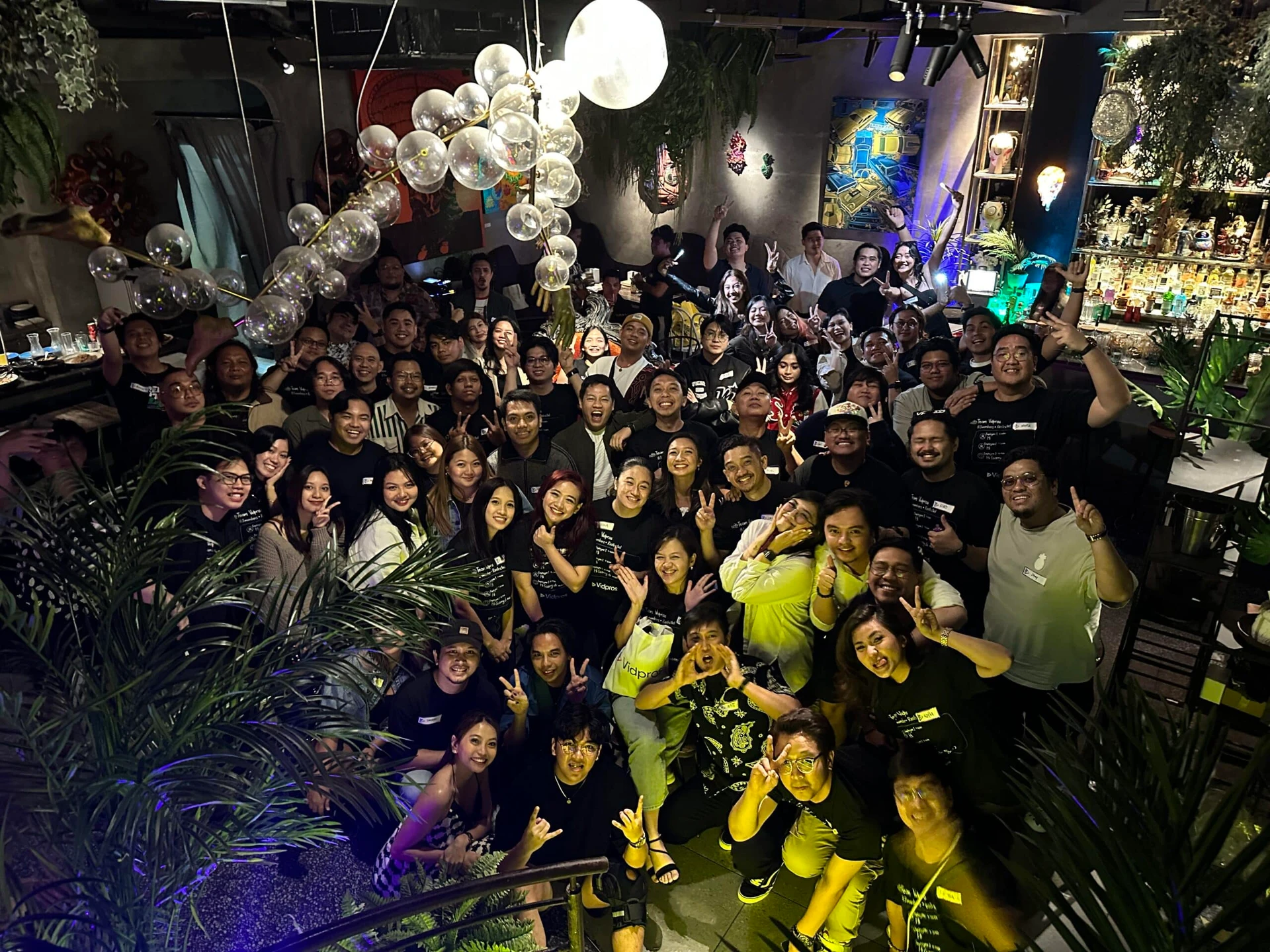 A large group of people gathered in a dimly lit venue celebrating with smiles and peace signs. Decorative balloons create an arch above the crowd. The setting includes lush plants and ambient lighting.