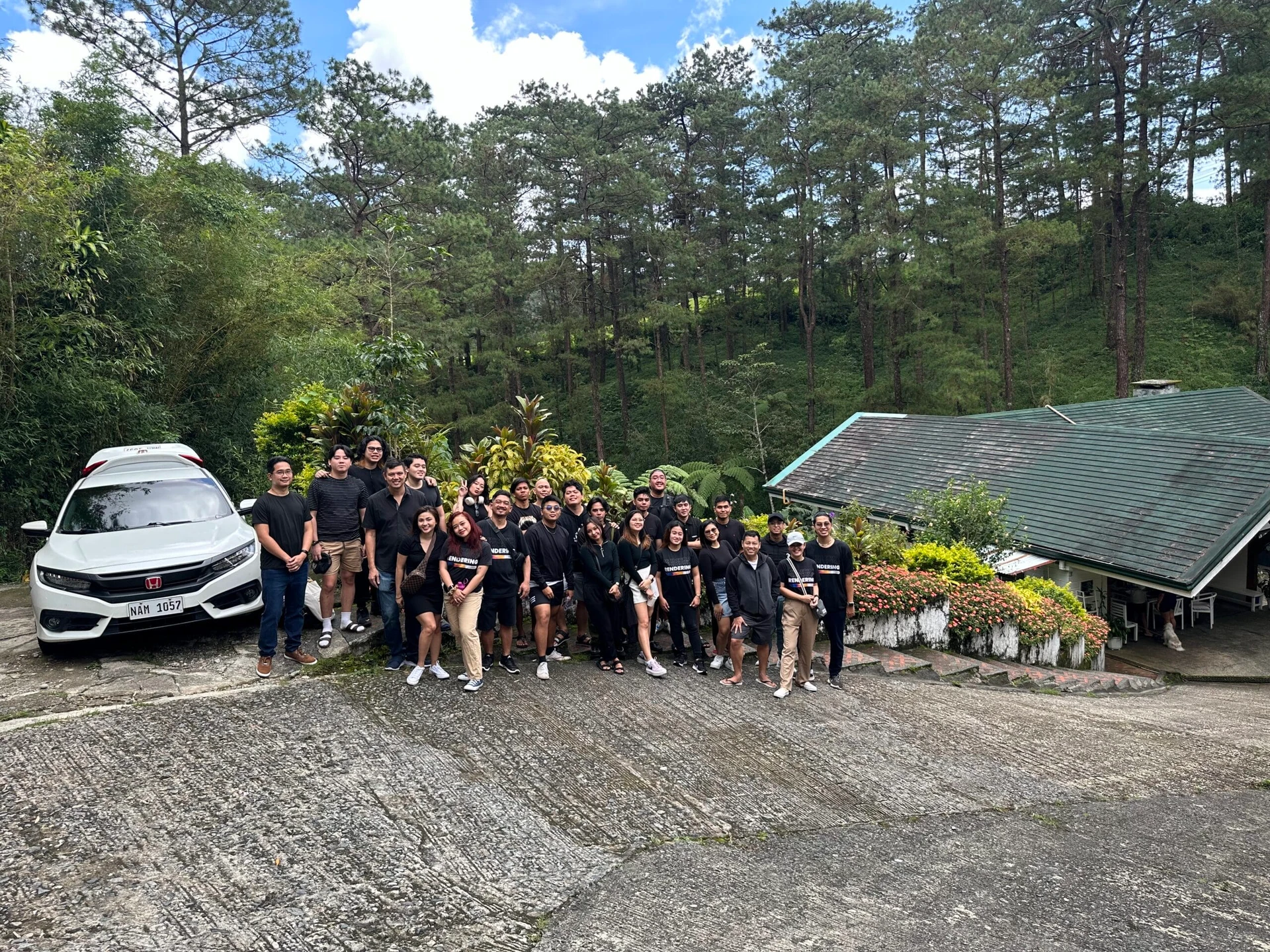 A large group of people pose together on a driveway beside a white car, in front of a house surrounded by lush greenery and tall pine trees. The sky is partly cloudy. Everyone is dressed casually.