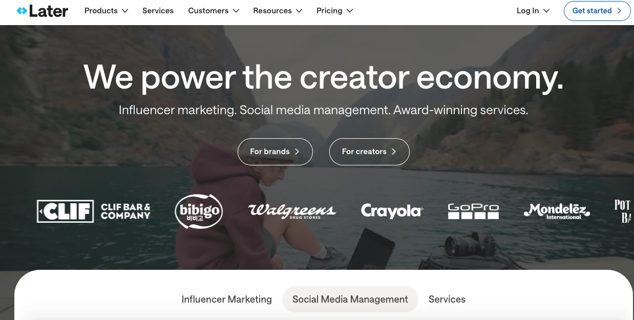 A person in a red jacket using a laptop by a lake with mountains in the background. Text reads, We power the creator economy. Influencer marketing. Social media management. Award-winning services. Logos of brands are displayed below.