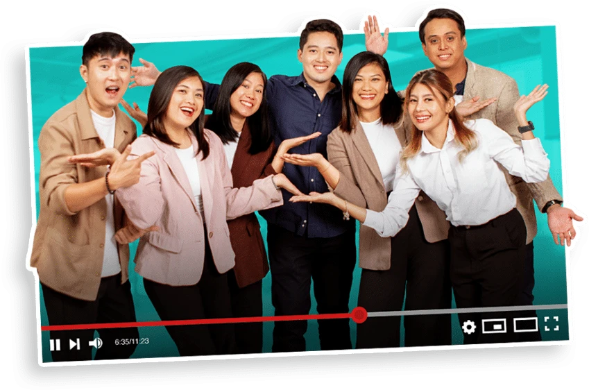 A group of seven people standing together, smiling and gesturing towards each other. They are wearing casual and semi-formal attire. The image has a video player overlay at the bottom with play and volume controls. The background is a solid teal color.