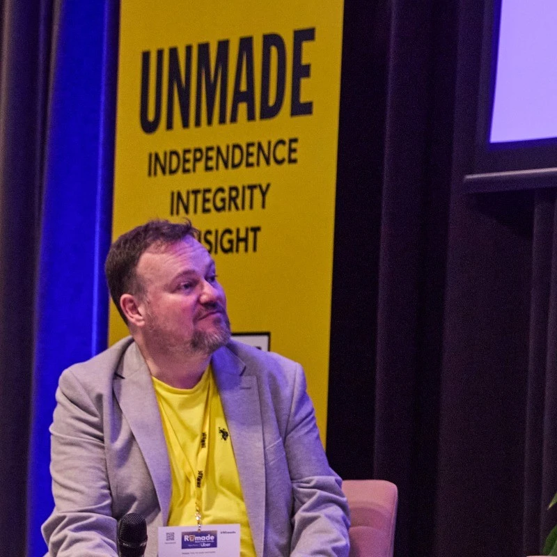 Man seated on a stage wearing a light gray blazer over a yellow shirt, holding a microphone. Behind him is a yellow banner with the words Unmade Independence Integrity Insight.