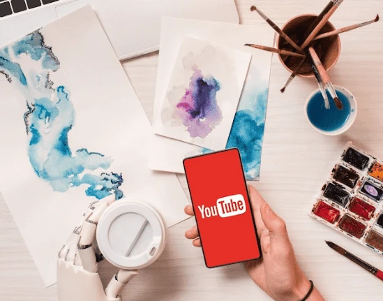 A robotic hand holds a smartphone displaying the YouTube logo, symbolizing the future of content creators. Nearby, a human hand holds a cup, while the table is scattered with watercolor paintings, brushes, and a palette. A laptop is partially visible in the top-left corner.